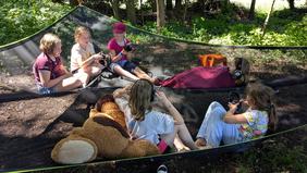 Kinder, auf Sommerfreizeit sitzen in einer Hängematte und hören mit Kopfhörern Musik.